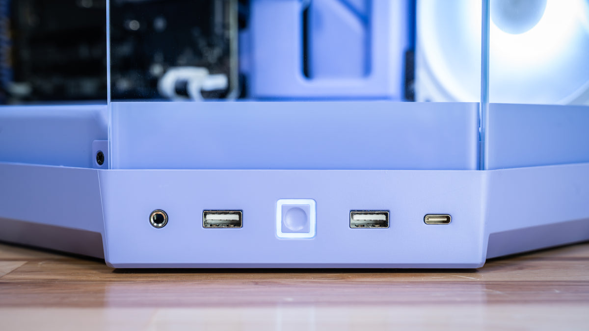 Close-up of the rear I/O panel of a light lavender-purple desktop PC case, showing a headphone jack, two USB-A ports, a USB-C port, and a central illuminated button, resting on a wooden surface.