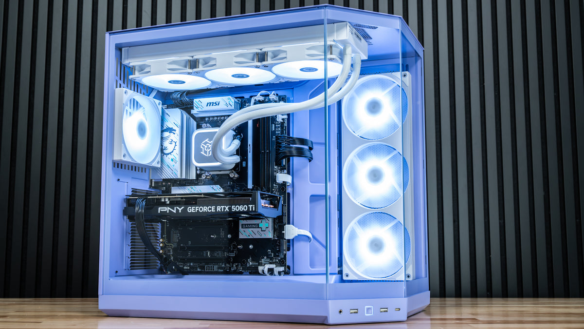 Custom gaming PC with transparent side panel, white RGB fans, liquid cooler, and PNY GeForce RTX 5060 Ti graphics card, on a wood surface against a dark paneled background