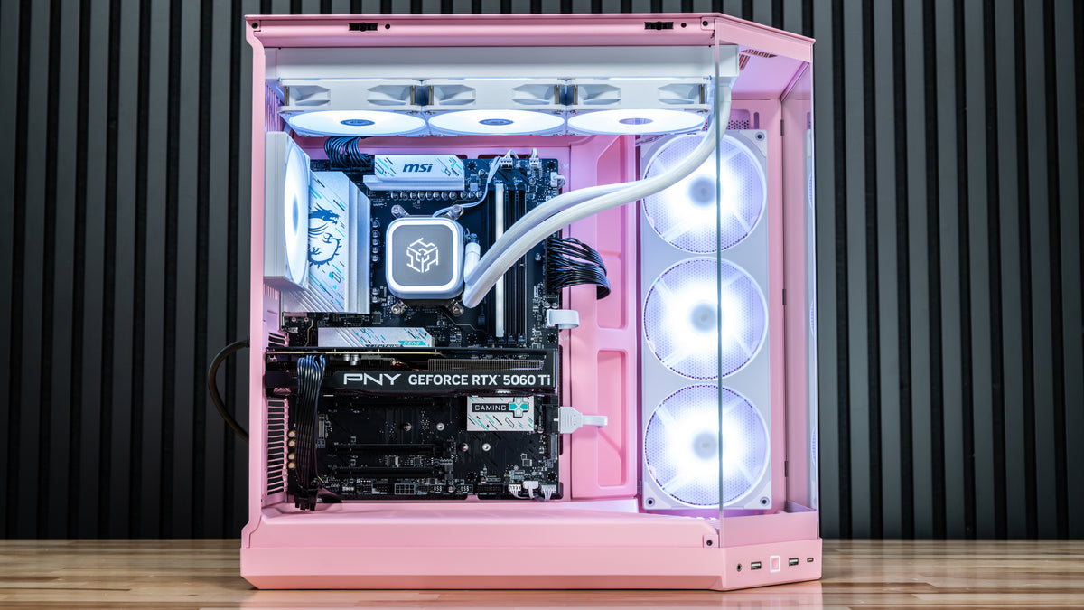 Pink custom-built desktop PC with a transparent side panel, showcasing a fully assembled interior with a liquid CPU cooler, PNY GeForce RTX 5060 Ti graphics card, and three rear-facing RGB fans, on a wooden surface against a dark slatted wall