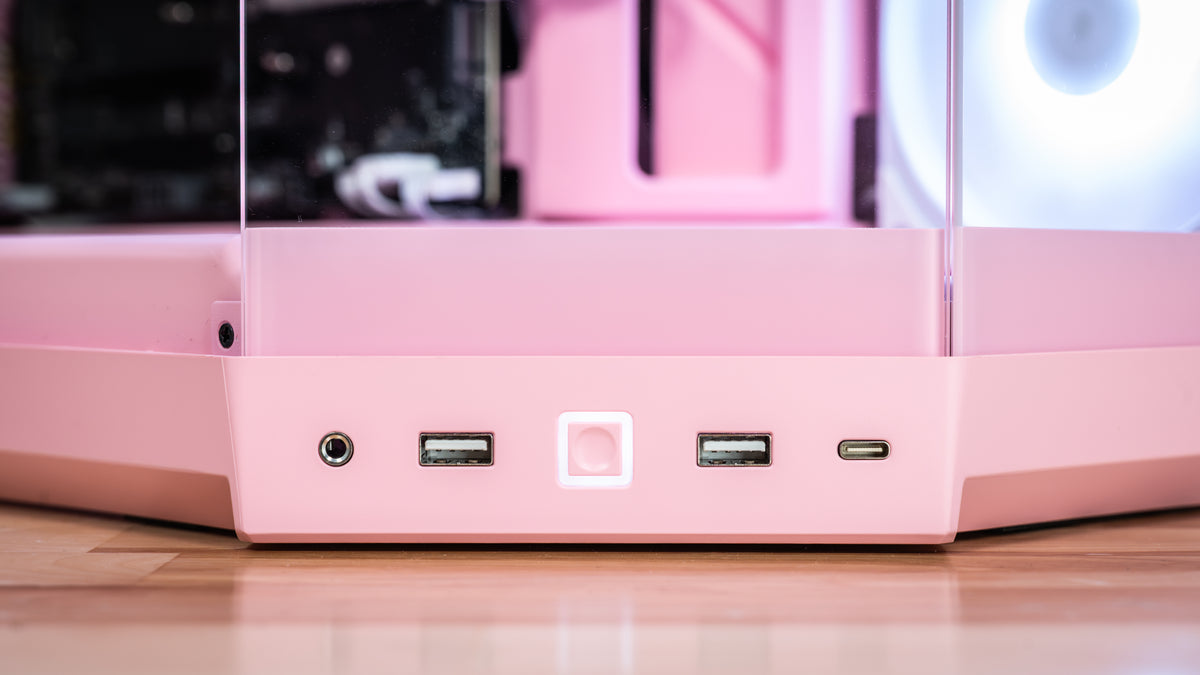 Close-up of the front I/O panel of a pink computer case, showing a headphone jack, two USB-A ports, a USB-C port, and a central power button with a glowing light, set on a wooden surface