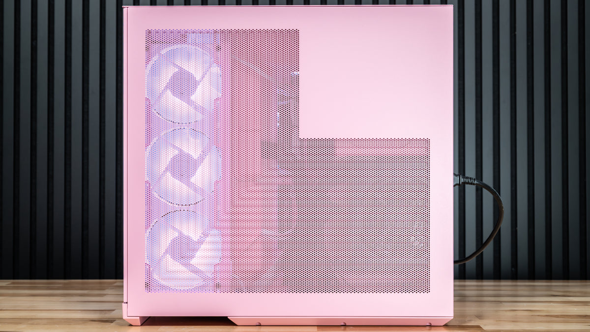 Pink computer case with mesh front panel and three visible fans, standing on a wooden floor against a dark slatted wall
